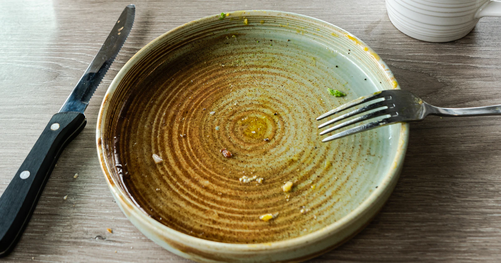 A finished meal on a table: an empty plate with a fork and knife, beside a coffee mug.